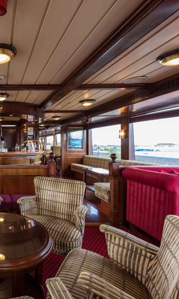 A classic ship lounge with wood-panelled walls, plush armchairs, a bar, and large windows overlooking a marina.