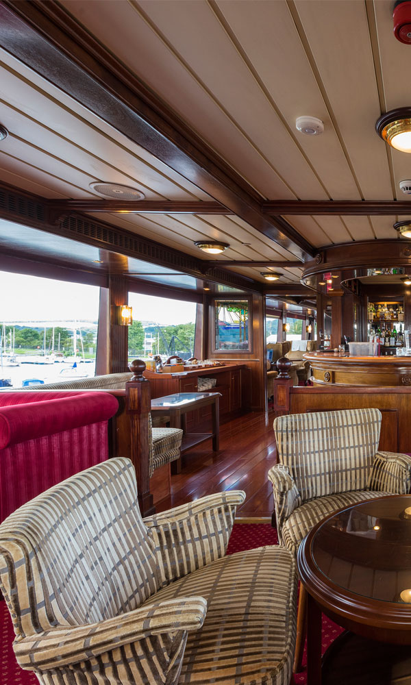 A classic ship lounge with wood-panelled walls, plush armchairs, a bar, and large windows overlooking a marina.